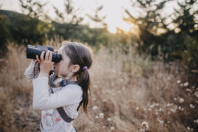 Side view of girl looking through binoculars while standing on field