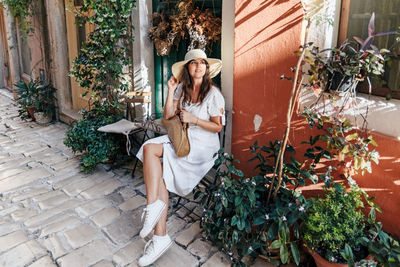 Portrait of an attractive young woman in summer dress sitting on chair in old town street.