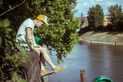 Side view of man sitting on retaining wall over river