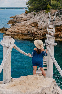 Rear view of young woman standing on old rope bridge over sea cove