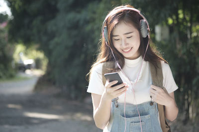 Smiling young woman listening music through headphones while standing against trees