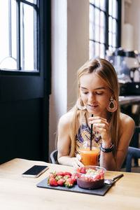 Pretty modern blonde female eating fresh strawberry while having healthy breakfast with berry smoothie bowl and fruit juice in cafe