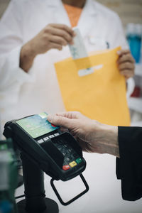 Cropped hand of senior female customer using contactless payment through credit card against pharmacist at checkout in s