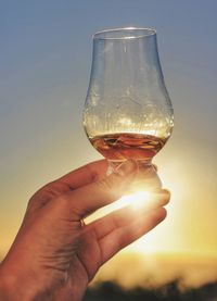 Close-up of hand holding wineglass against sky during sunset
