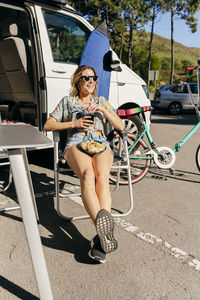 Cheerful woman enjoying a juice while sitting out of caravan in camping near surf board and bicycle