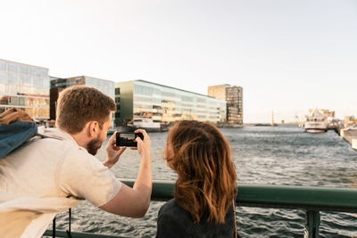 Woman looking at man photographing river on smart phone while traveling in city