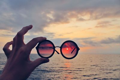 Cropped hand holding sunglasses at beach during sunset