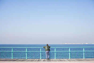 Rear view of man standing on railing against sea and sky