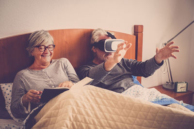 Woman smiling while senior man looking through virtual reality simulator on bed at home