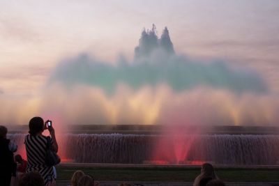Rear view of woman photographing colorful fountain during sunset