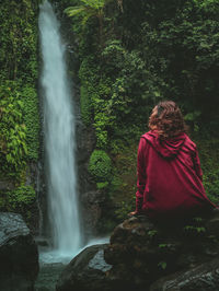 Rear view of woman looking at waterfall while sitting in forest