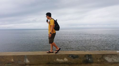 Side view of young man walking on retaining wall by sea against sky