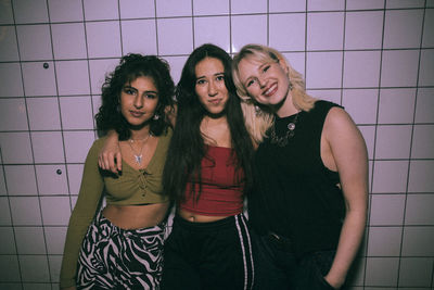 Portrait of young multiracial female friends standing with arms around against white wall at nightclub