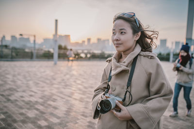 Thoughtful woman looking away while holding digital camera on street in city during sunset