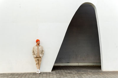 Full length thoughtful asian male wearing casual clothes and orange hat leaning with hands in pockets on white building wall with creative arch