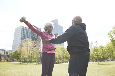 Man teaching dumbbell exercise to senior woman at public park