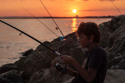 Side view of young man photographing sea against sky during sunset