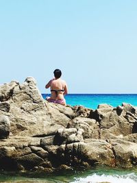 Rear view of shirtless man on rock in sea against clear sky