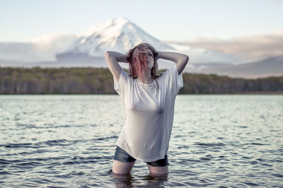 Young woman with hands behind head standing in lake against sky during sunset