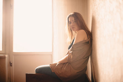 Portrait of young woman sitting on headboard of bed by window at home