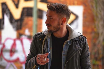 Young man looking away while holding cigarette against graffiti wall