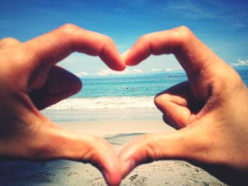 Cropped image of hands holding heart shape on beach against sky