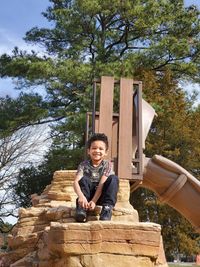 Portrait of boy sitting on wood against trees