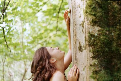 Close-up of woman looking up while standing by wall against trees