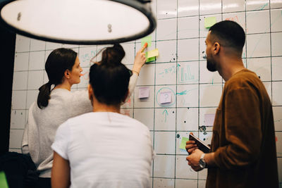 Young female computer programmer explaining strategy on wall to multi-ethnic colleagues at small creative office