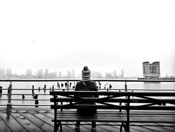 Rear view of woman wearing warm clothing while sitting on bench by river in city against clear sky
