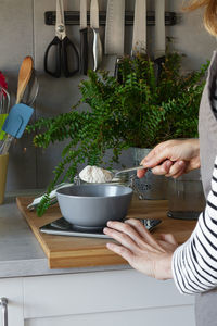 Crop hands of faceless woman in apron adding spoon of flour into bowl on scale while cooking in modern kitchen