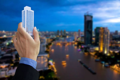 Cropped hand of businessman holding model over cityscape against cloudy sky at dusk