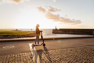 Full length of woman riding electric push scooter on road by sea against sky during sunset