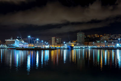 Illuminated buildings by sea against sky at night