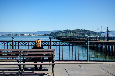 Rear view of woman sitting on bench by river against clear blue sky