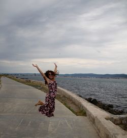 Excited woman jumping over pier by sea against cloudy sky