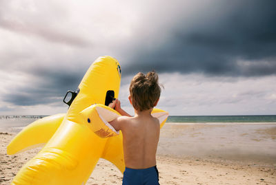 Rear view of shirtless boy playing at beach against cloudy sky