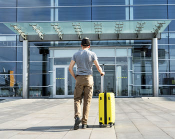 Rear view of young man traveler in casual carrying yellow suitcase next to entrance to airport 