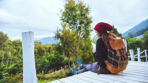 Rear view of woman wearing knit hat sitting on plank against sky