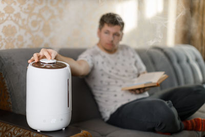Young caucasian man hold book in hand and change direction of air flow of white modern humidifier