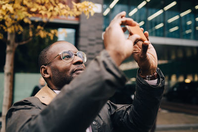 Businessman photographing with using smart phone while standing against modern building in city