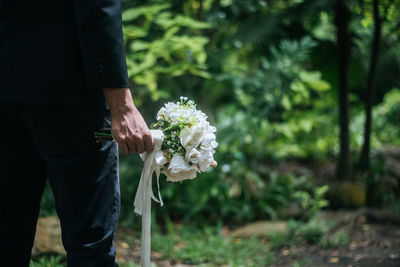 Midsection of groom holding flower bouquet while standing against trees in forest