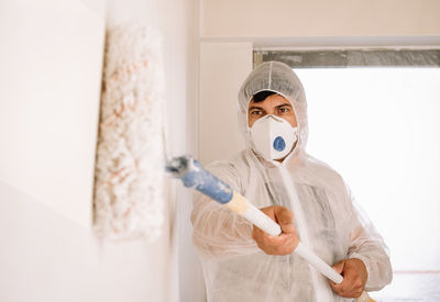 Man in protective workwear and mask working and holding paint roller against white wall at home