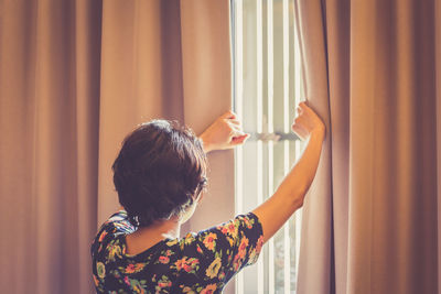 Rear view of mature woman looking through window at home