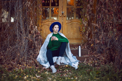 Woman in green sweater and plaid sitting on porch,house with ivy in autumn drinking tea from thermos