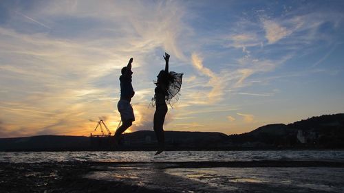 Silhouette couple jumping at beach against sky during sunset