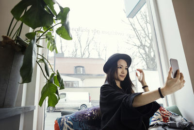 Smiling young woman taking selfie while sitting against window in office