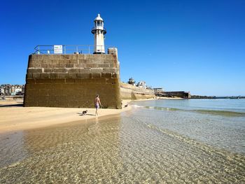 Lighthouse by sea against clear blue sky