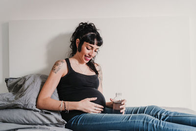 Smiling pregnant woman with water bottle sitting on bed at home