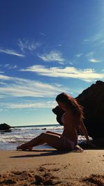 Side view of young woman sitting at beach against sky during summer